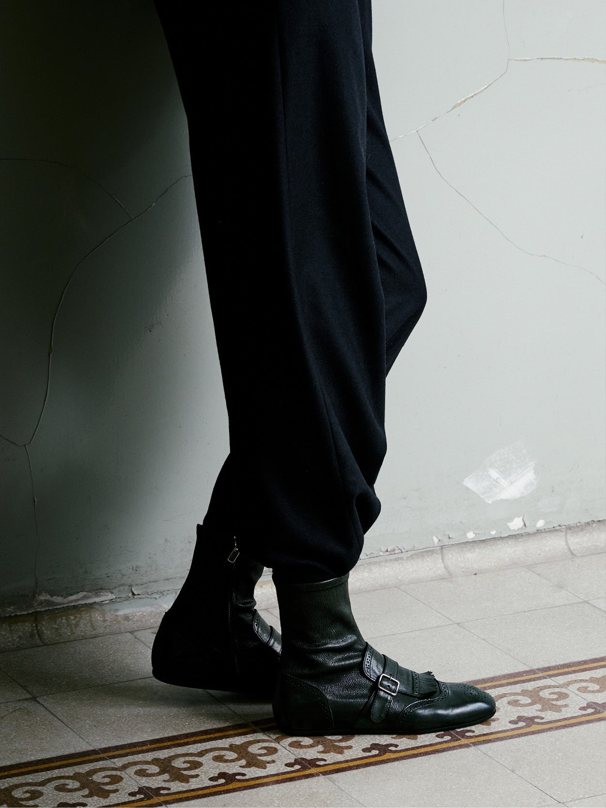 A close-up of a person's legs wearing black wide-leg trousers and black leather buckle-front ankle boots, standing on a patterned tile floor against a textured light gray wall.