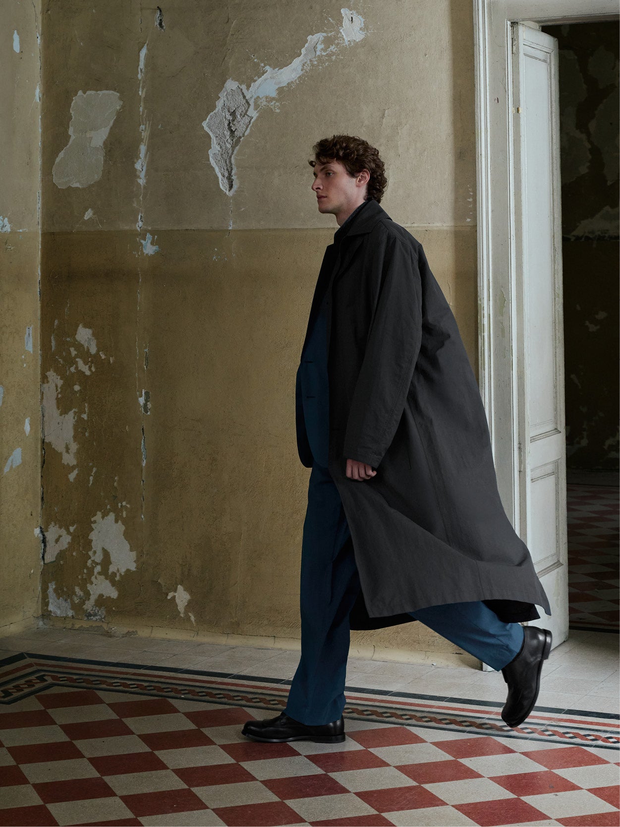A man in a long dark charcoal coat, blue trousers, and black shoes walks in profile through a doorway in a building with peeling beige walls and a red-and-white checkered floor.