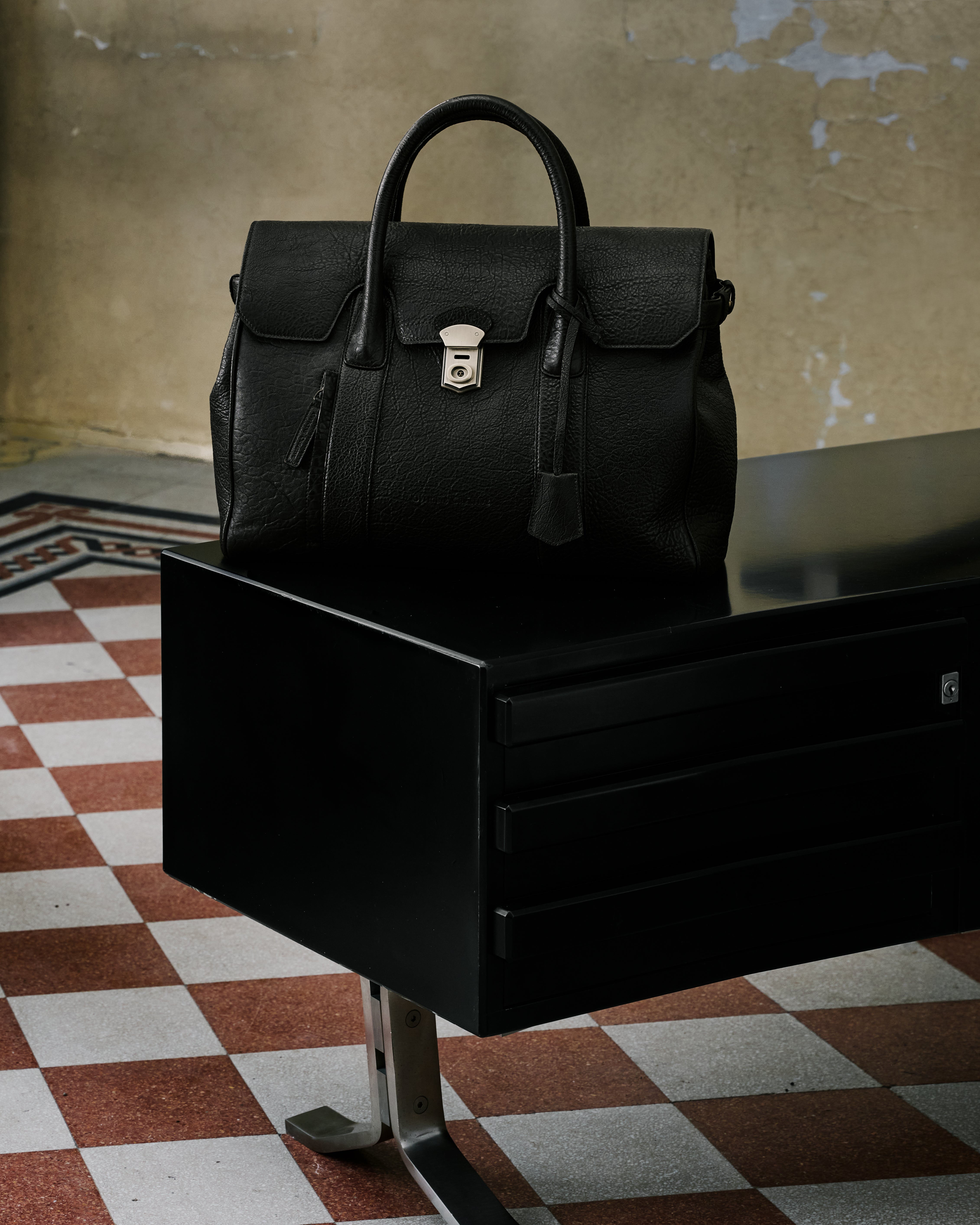 A black leather briefcase with a silver-tone turn-lock clasp and top handle, displayed on a black sideboard against a textured beige wall and checkered floor.