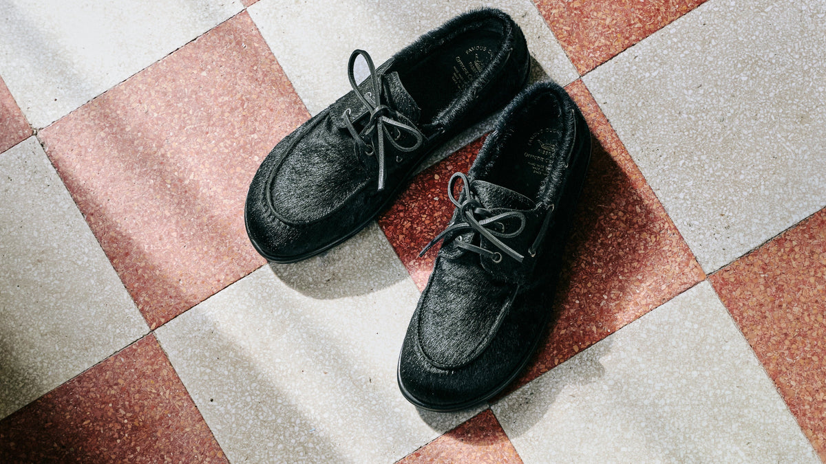 A pair of black pony boat shoes photographed from above on a "cementine" style floor with pink, white, and gray checkered tiles.