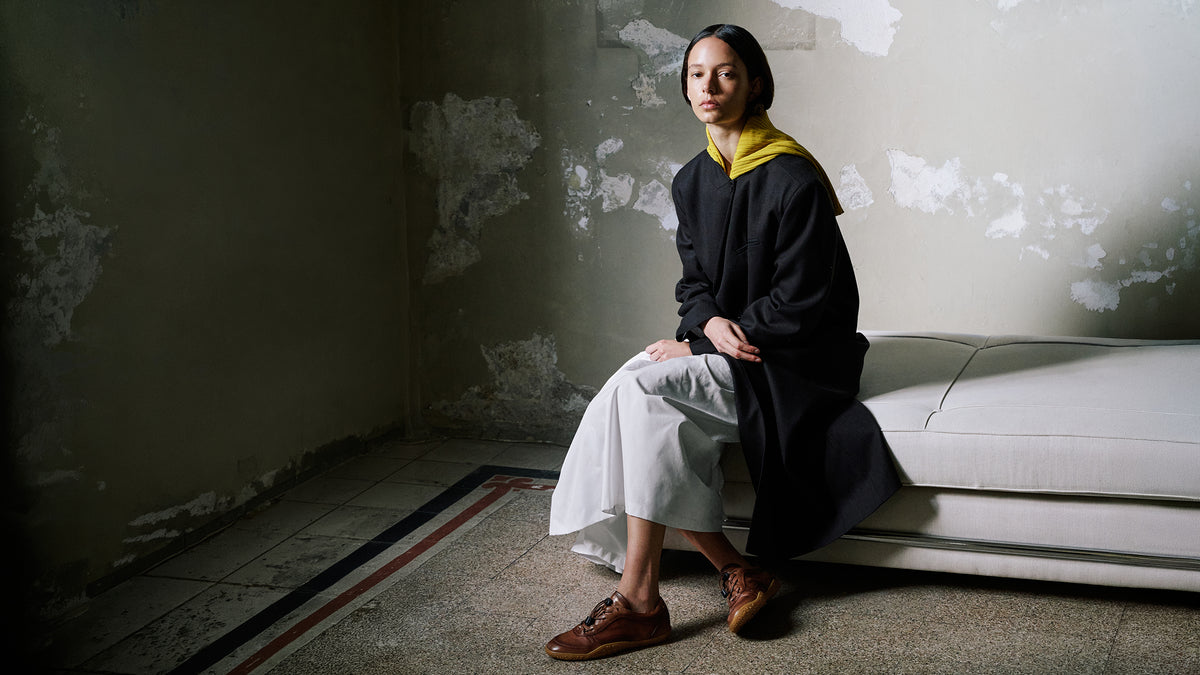 A woman sits on a white bench in a dimly lit room with peeling plaster walls. She is wearing a black long-sleeve top with a yellow collar, light gray wide-leg pants, and brown leather shoes.